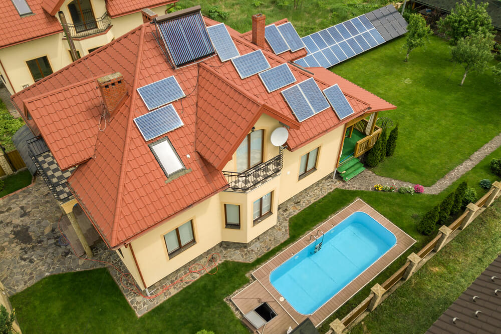 Aerial View of a New Autonomous House With Solar Panels and Water Heating Radiators on the Roof and Green Yard With Blue Swimming Pool.