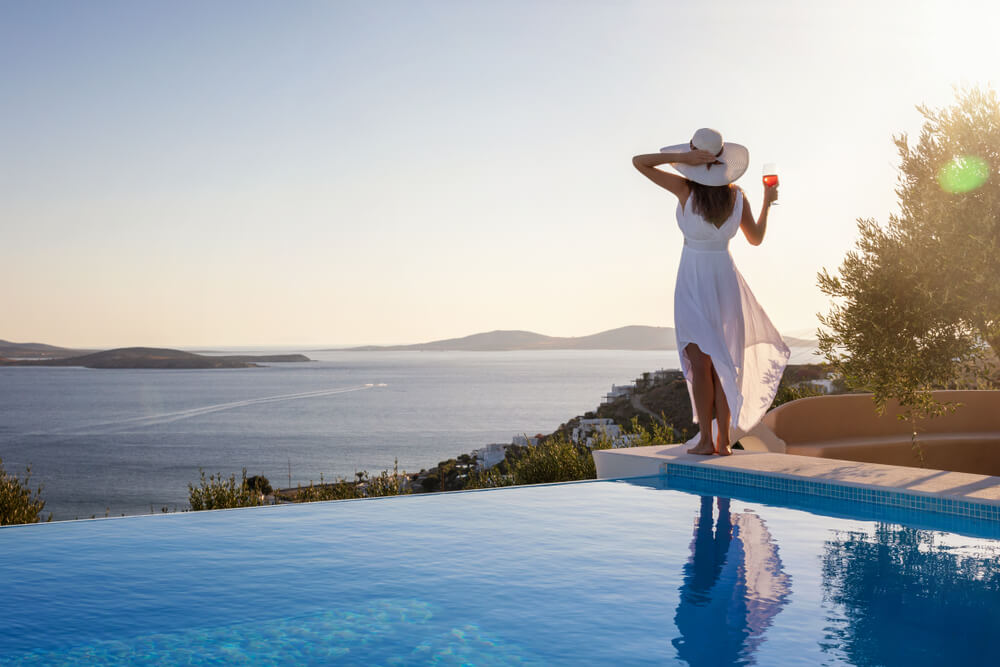 Beautiful Woman in White Dress and Hat Stands by the Pool Edge and Enjoys the Summer Sunset With a Glass of Wine and View to the Mediterranean Sea