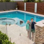 Two young children in swimwear standing beside a glass security gate at a backyard swimming pool, showcasing modern frameless Pool Fence Ideas that combine safety with unobstructed views.